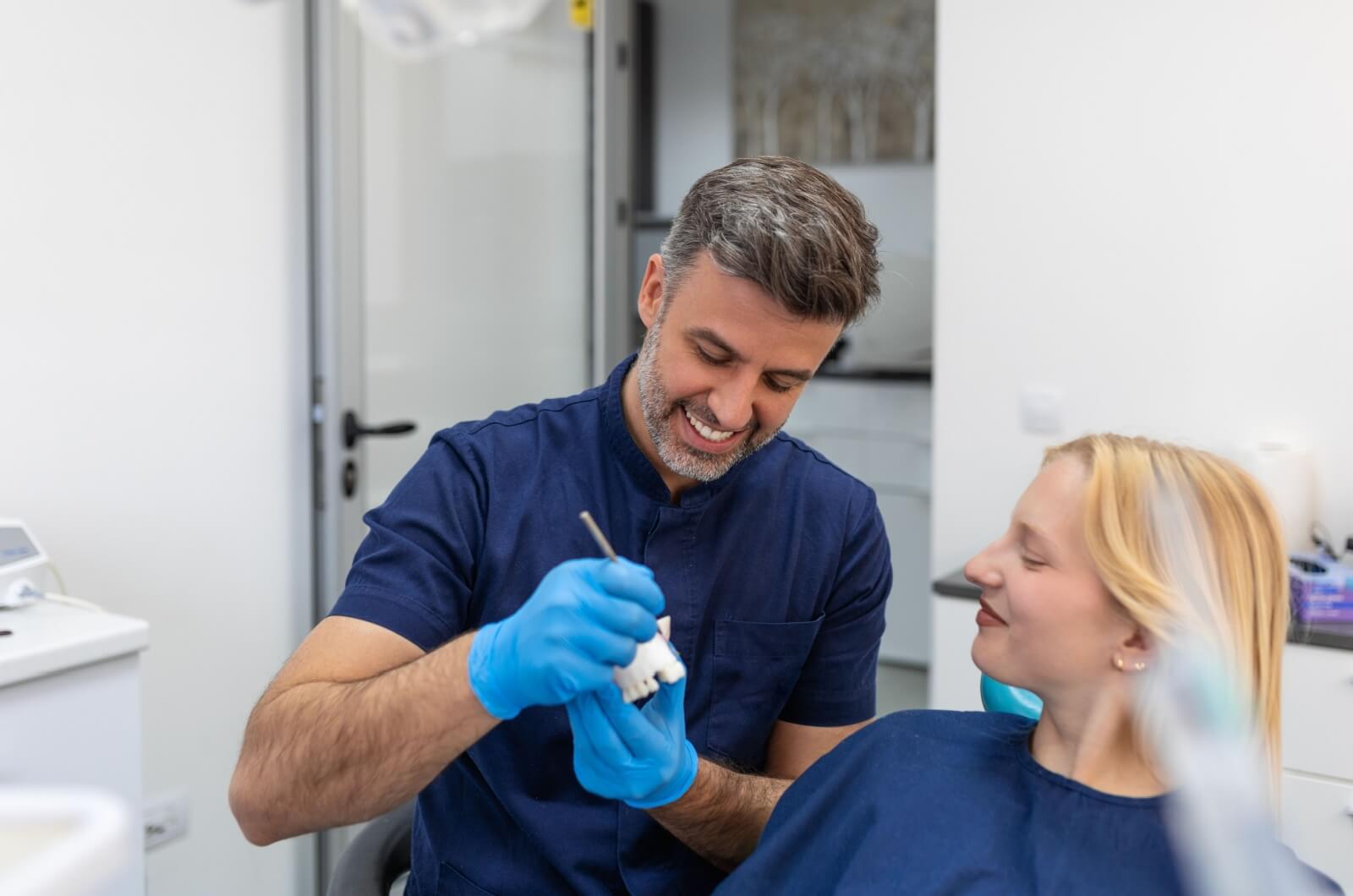 A smiling dentist shows a patient on a dental plaster mold where her dental bridge is in her mouth and how it can be repaired