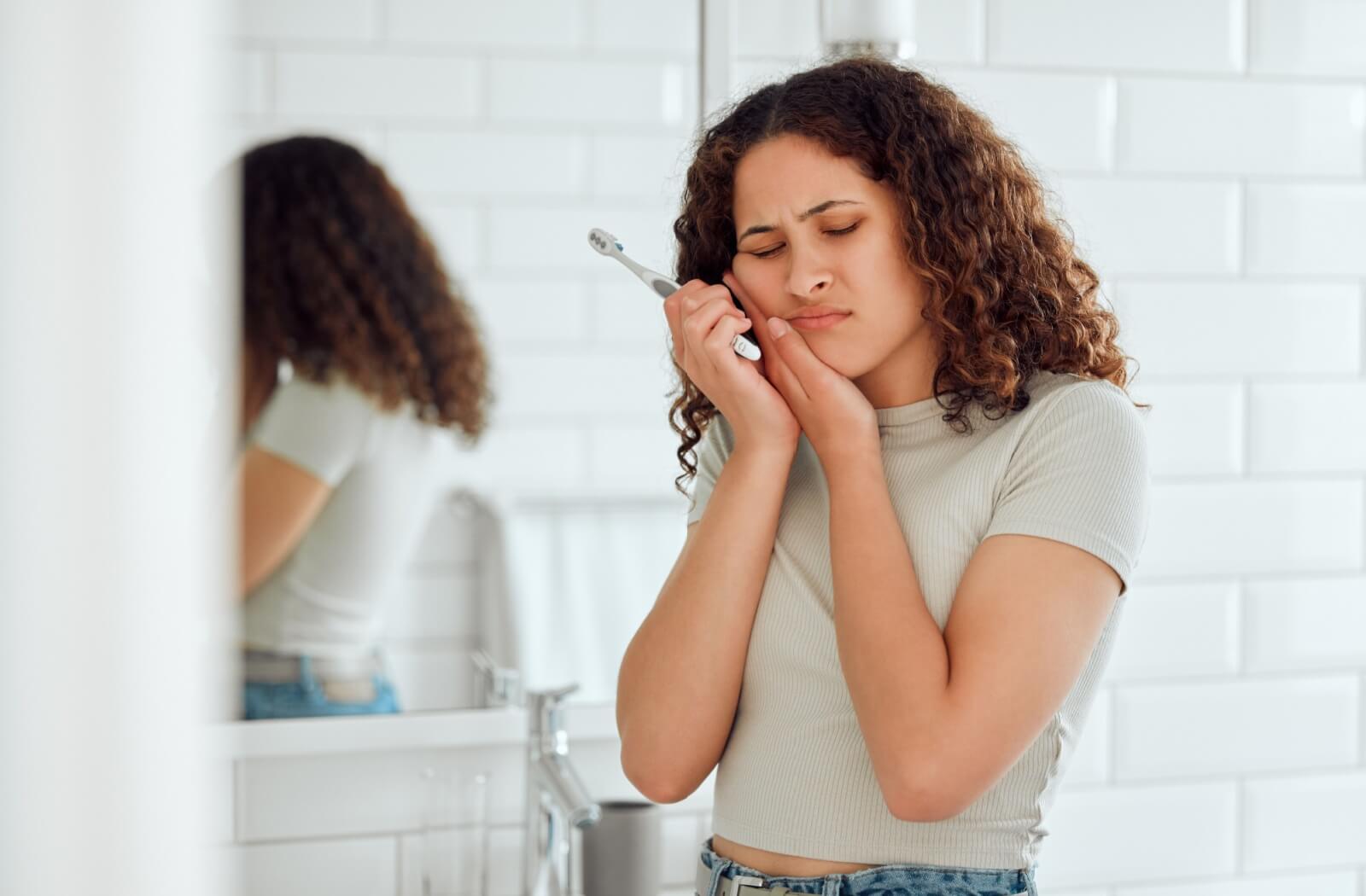 A young woman clutches her face in pain while holding her toothbrush.