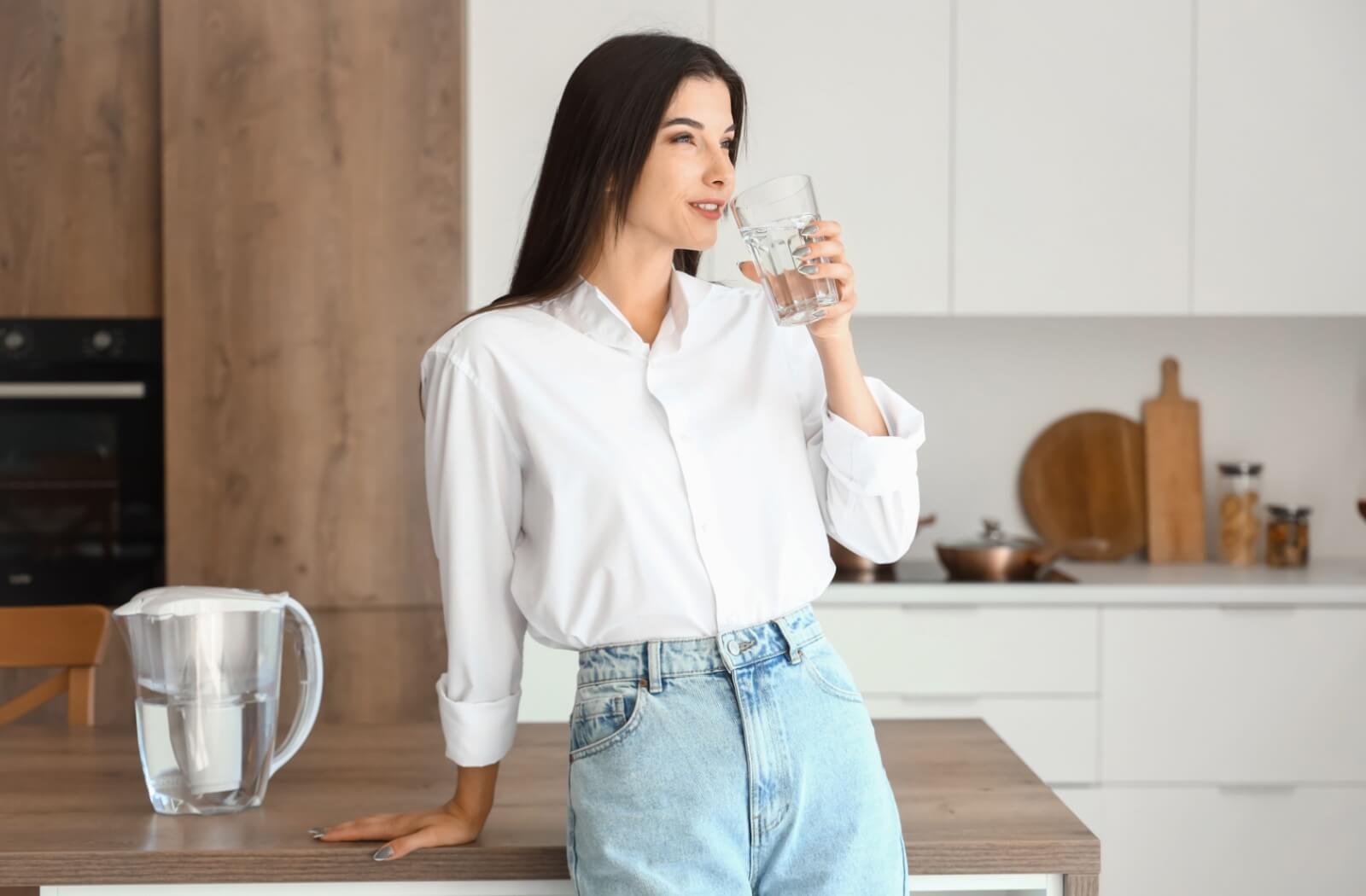 A happy young woman sits in her kitchen drinking a glass of water with a jug of seltzer water on the table beside her